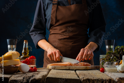 The chef cuts the chicken fillet on a cutting board. Lots of ingredients. Wooden texture. Restaurant  hotel  home cooking. Dark background. Menu design. Close-up.