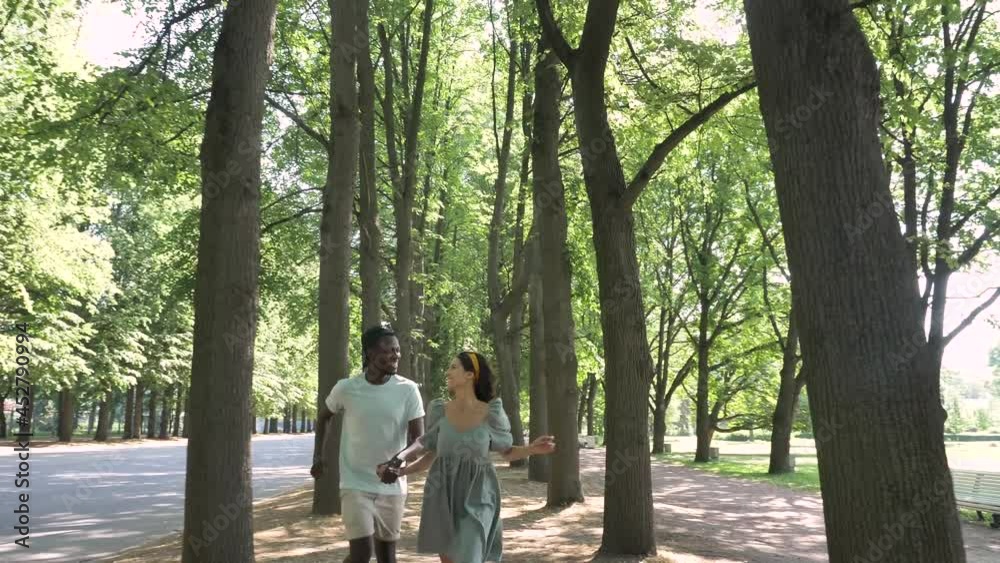 African American man and brunette woman run joining hands and laugh among trees casting shadows in city park on sunny summer day African American man and brunette woman run joining hands and laugh among trees casting shadows in city park on sunny summer day
