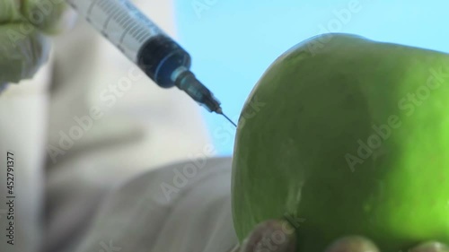 Close-up of a human in a medical coat and gloves injecting a syringe into an apple with some liquid on a blue background