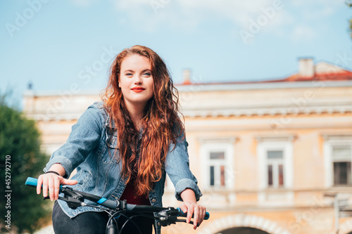 Half-length portrait of long red curly hair caucasian teenager girl riding a modern bicycle in the old city center. Diverse people beauty or city bike hiring concept image.