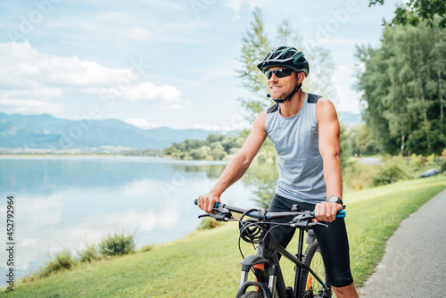 Konstfotografi Portrait of a sincerely smiling man dressed in cycling clothes, helmet and sunglasses riding a bicycle on the asphalt out-of-town bicycle path along a mount lake