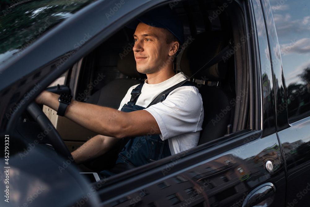 Obraz premium In the form of a courier, a man of Caucasian appearance works at the wheel of a car. The transport company driver delivers the goods. The entrepreneur is young in his business.