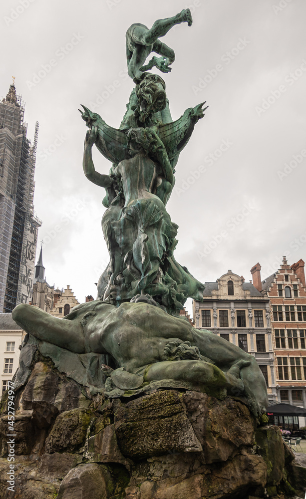 Antwerpen, Belgium - August 1, 2021: Green bronze Brabo statue on Grote Markt. Closeup of defeated Antigoon body with view up onto Brabo against gray sky. Buildings as backdrop.