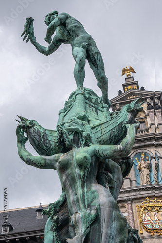 Antwerpen, Belgium - August 1, 2021: Green bronze Brabo statue on Grote Markt. Detail about throwing the hand, against gray sky. Gable of city hall in back.