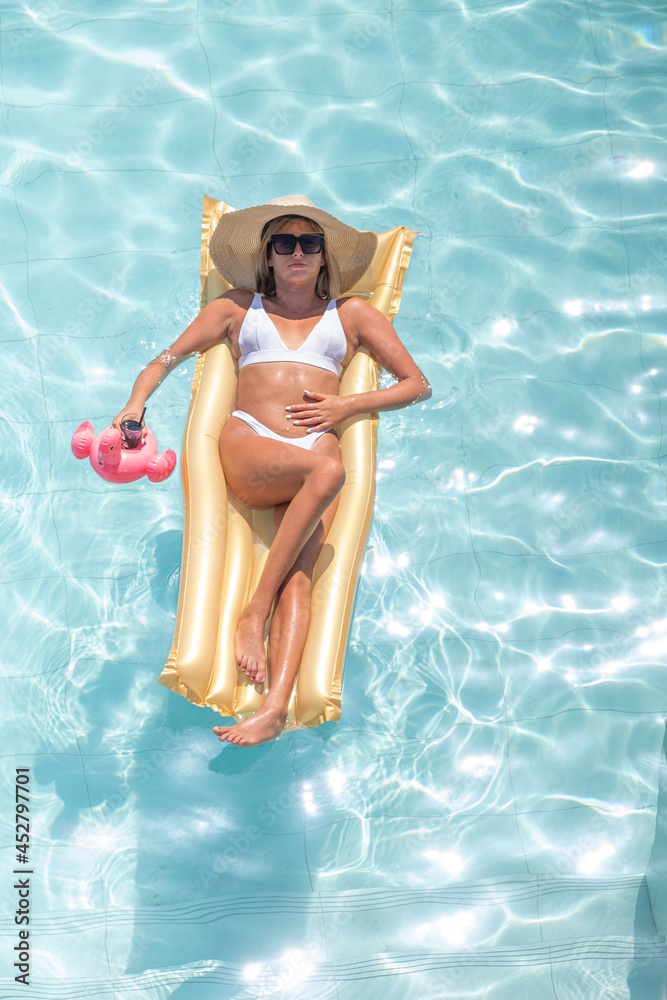 Top view of girl in a bathing suit and hat floating on inflatable gold mattress in the pool with drink cocktail. Summer holidays and relax concept