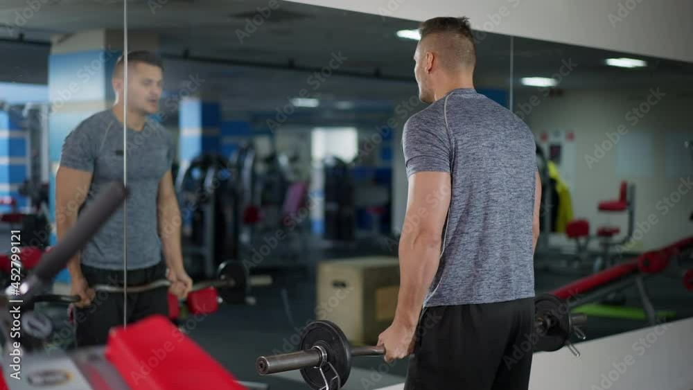 Back view of strong sportsman lifting heavy barbell reflecting in mirror in gym. Confident motivated Caucasian man working out training indoors. Weightlifting and lifestyle concept Back view of strong sportsman lifting heavy barbell reflecting in mirror in gym. Confident motivated Caucasian man working out training indoors. Weightlifting and lifestyle concept