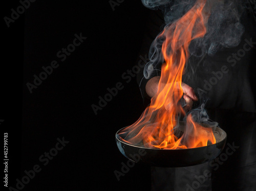Obraz na plátně Professional chef cooking food in pan with fire flame on black background