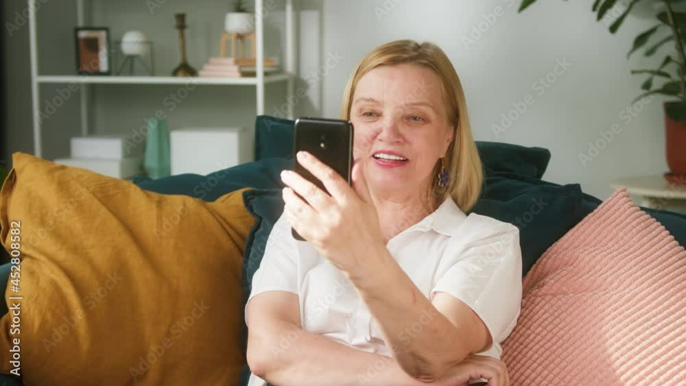 Elderly woman sitting on sofa in living room and speaking on video call using smartphone. Female pensioner communicating online with children. Smiling grandmother during leisure time at home Elderly woman sitting on sofa in living room and speaking on video call using smartphone. Female pensioner communicating online with children. Smiling grandmother during leisure time at home