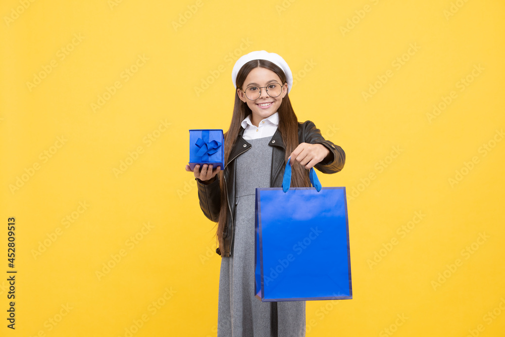 Happy teenage girl back to school holding gift box and paperbag yellow background, shopping bag