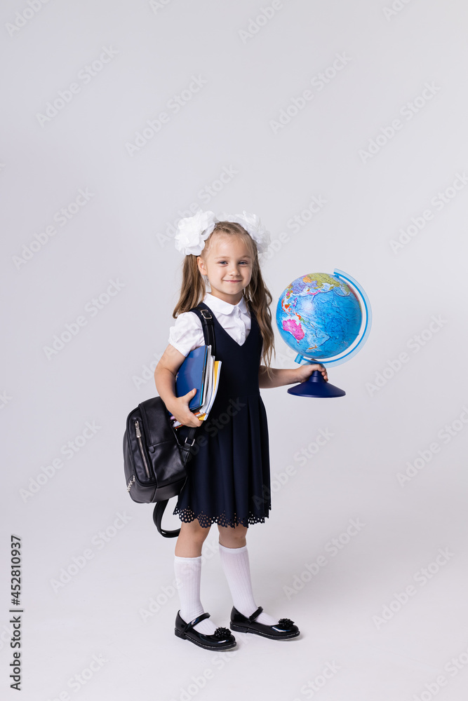 Fototapeta premium Elegant European schoolgirl holds a globe, books and a briefcase. A girl with white bows on a white background. The student smiles.