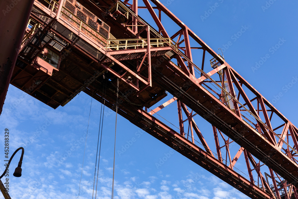 Massive overhead industrial structure with open stairs and open grates against a blue sky, horizontal aspect