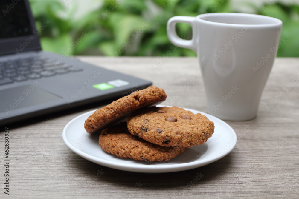 Pile of three delicious cookies on white plate. Blurry white glass and laptop on the background Pile of three delicious cookies on white plate. Blurry white glass and laptop on the background