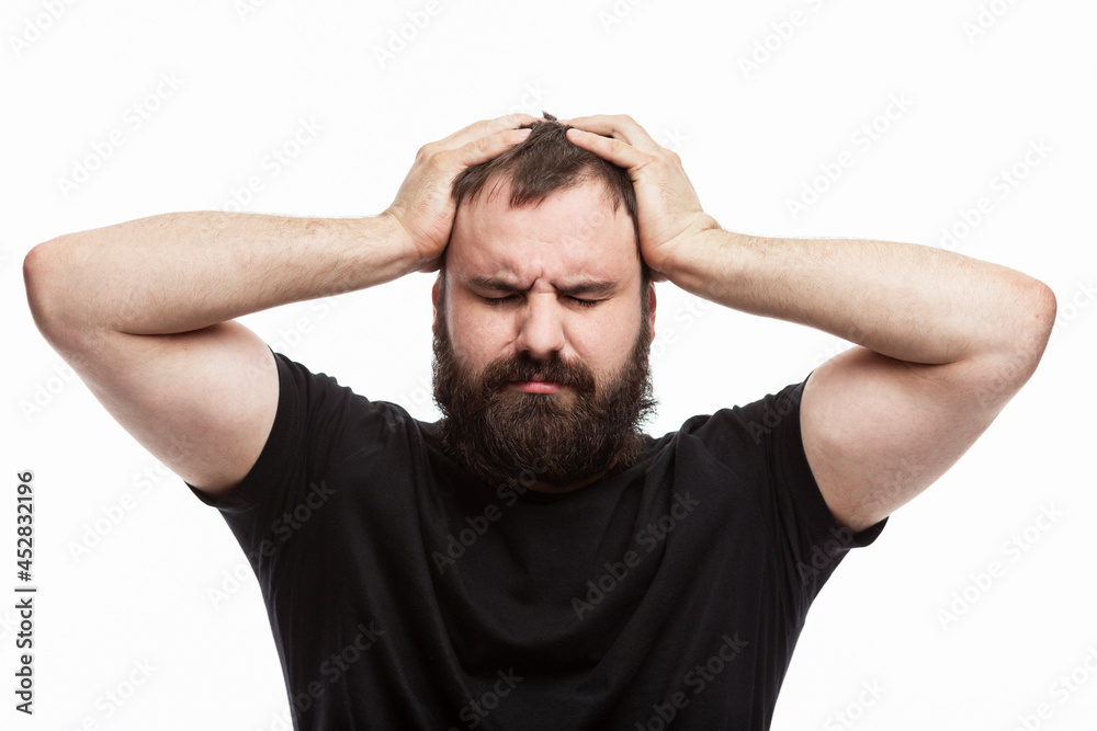 A man with a beard is emotionally holding his head. Depression and stress. White background. Brunette in a black T-shirt. A man with a beard is emotionally holding his head. Depression and stress. White background. Brunette in a black T-shirt.