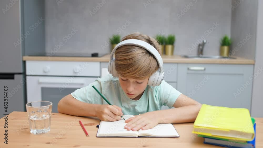 Distance learning, caucasian teen boy sitting at workplace in living room and uses a laptop to study in self-isolation at home, homeschooling online by videocall, the boy writes in a notebook. Distance learning, caucasian teen boy sitting at workplace in living room and uses a laptop to study in self-isolation at home, homeschooling online by videocall, the boy writes in a notebook.