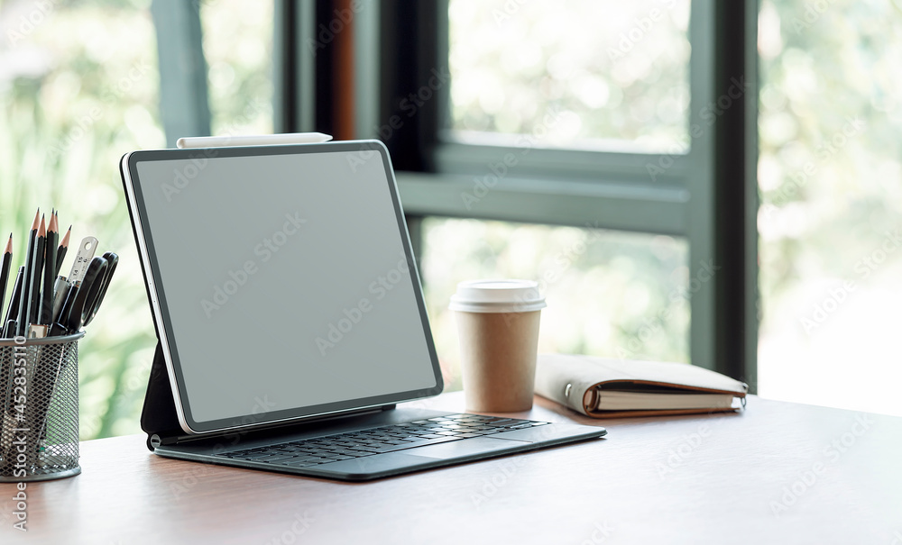 Fototapeta premium Blank screen tablet with magic keyboard and supplies on wooden table in modern office room.