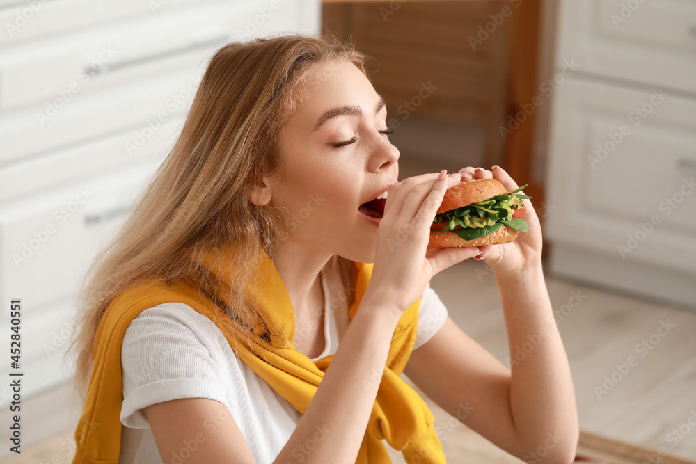 Young woman eating tasty vegan burger in kitchen Young woman eating tasty vegan burger in kitchen