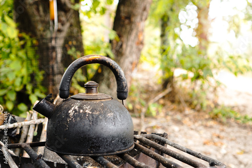 Old metal tea pot covered with soot and dust on grill. Making tea in nature camping in the forest. Cooking on fire in summer.