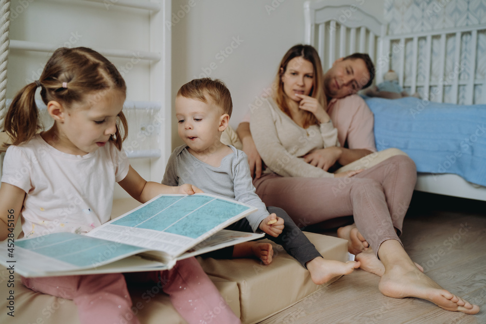 cute little boy and a boy reading book in child room. Young parents on background sitting on the floor by the cot cute little boy and a boy reading book in child room. Young parents on background sitting on the floor by the cot