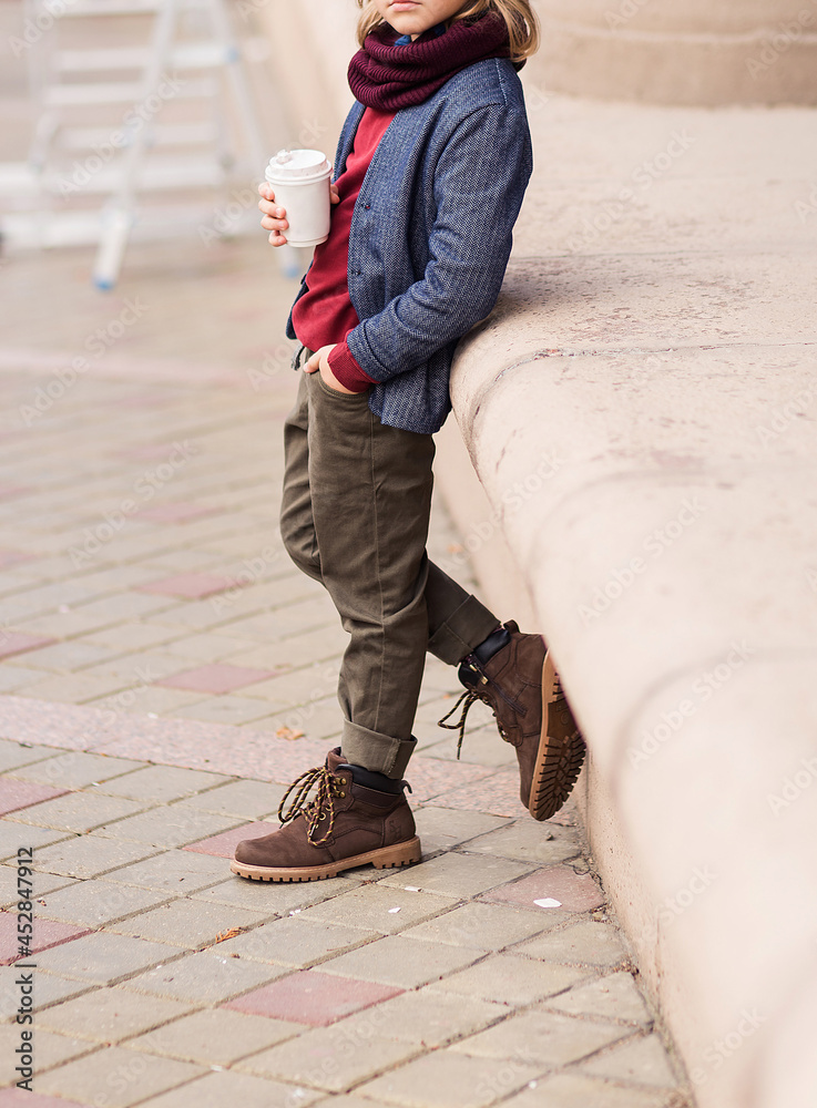 Stylish boy in trousers, a jacket and a scarf posing on a city street with a glass of hot drink in his hand Stylish boy in trousers, a jacket and a scarf posing on a city street with a glass of hot drink in his hand