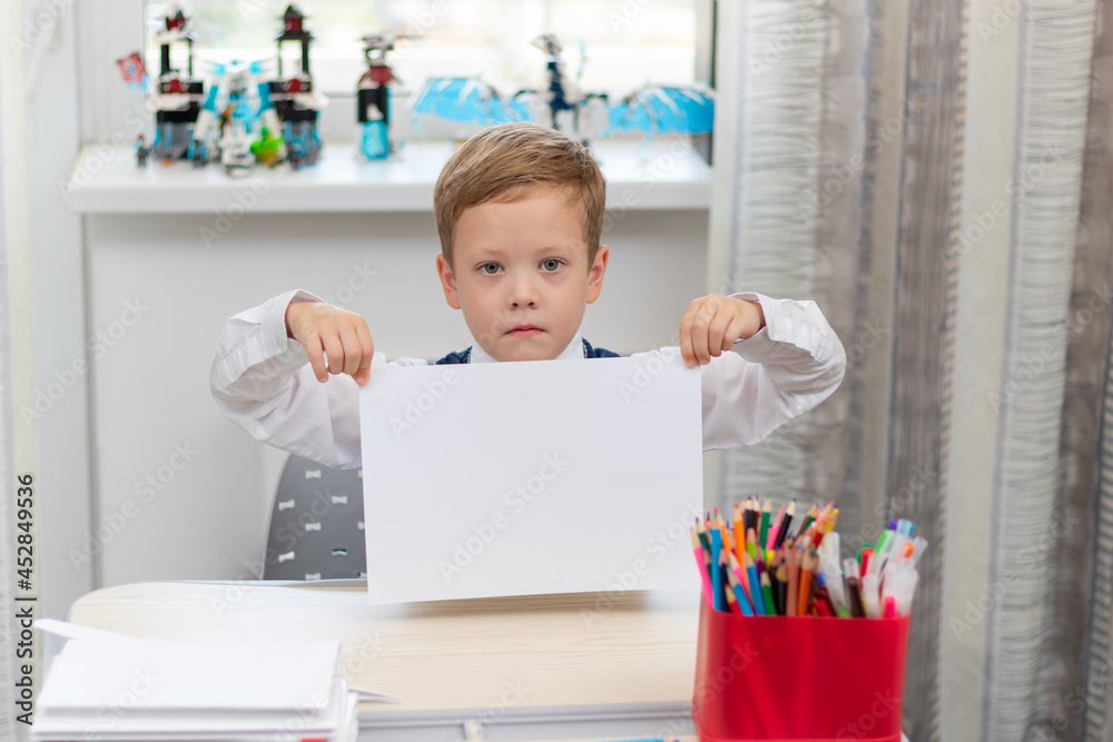 A cute boy first grader in a school uniform at home while isolated at his desk holds a sheet of blank paper. Selective focus. Portrait A cute boy first grader in a school uniform at home while isolated at his desk holds a sheet of blank paper. Selective focus. Portrait
