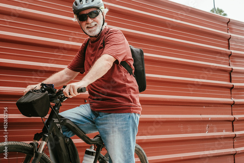Senior cyclist in outdoor excursion with electric bicycle resting against a red metal panel looking at camera