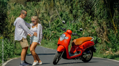 Two caucasian tourist woman man dance near scooter. Love couple on red motorbike in white clothes, sunglasses on forest road trail trip. Dancing road. Motorcycle rent. Asia Thailand ride tourism.