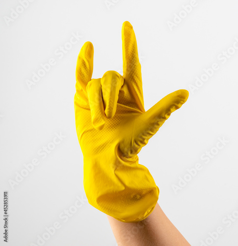 A hand in yellow rubber gloves shows a rock horn, two fingers up on a white background. Woman's hand in yellow rubber gloves shows 'Horns Up' sighn