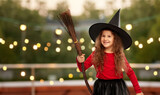 halloween, holiday and childhood concept - smiling girl in costume and witch hat with broom over garland lights at roof top party background