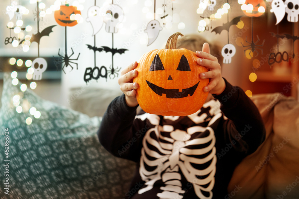 Naklejka premium halloween, holiday and childhood concept - smiling little boy in costume of skeleton with jack-o-lantern sitting on sofa at home