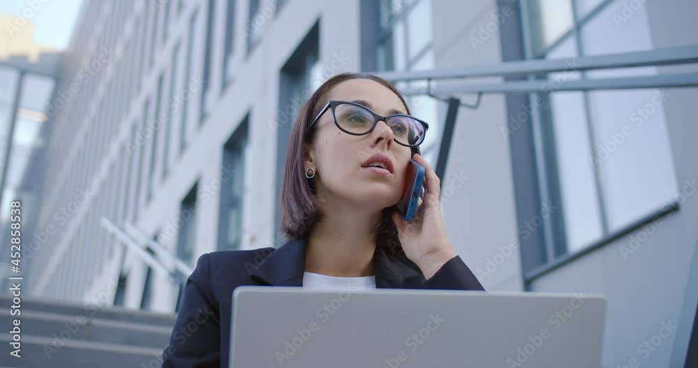 Close up of Caucasian young busy female executive manager sitting on stairs outdoors near office center working on laptop and talking on smartphone. Businesswoman calling on cellphone, job concept Close up of Caucasian young busy female executive manager sitting on stairs outdoors near office center working on laptop and talking on smartphone. Businesswoman calling on cellphone, job concept