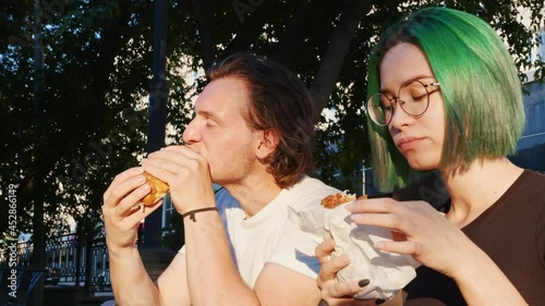 Guy and girl eat fastfood on city street. Quick bite, snack, takeaway, takeout, carry-out food. Hungry young man, woman chew hamburgers with pleasure. Unhealthy junk products addiction