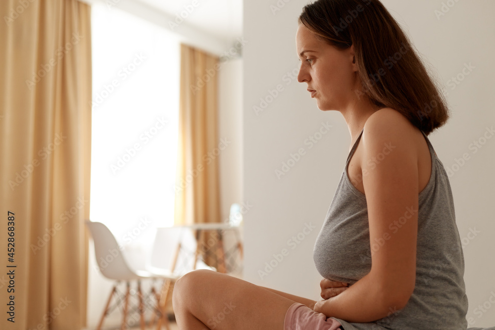 Side vie portrait of ill famale suffering from stomachache, woman wearing gray sleeveless t shirt keeping hands on sick belly, suffering from pain during period. Side vie portrait of ill famale suffering from stomachache, woman wearing gray sleeveless t shirt keeping hands on sick belly, suffering from pain during period.