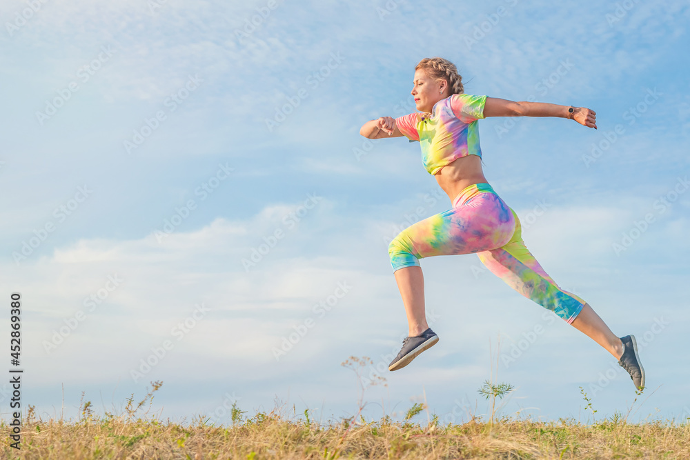 Young woman in a bright light tracksuit flies in a long stride over the meadow against the background of the blue summer sky. Outdoor sports. Young woman in a bright light tracksuit flies in a long stride over the meadow against the background of the blue summer sky. Outdoor sports.