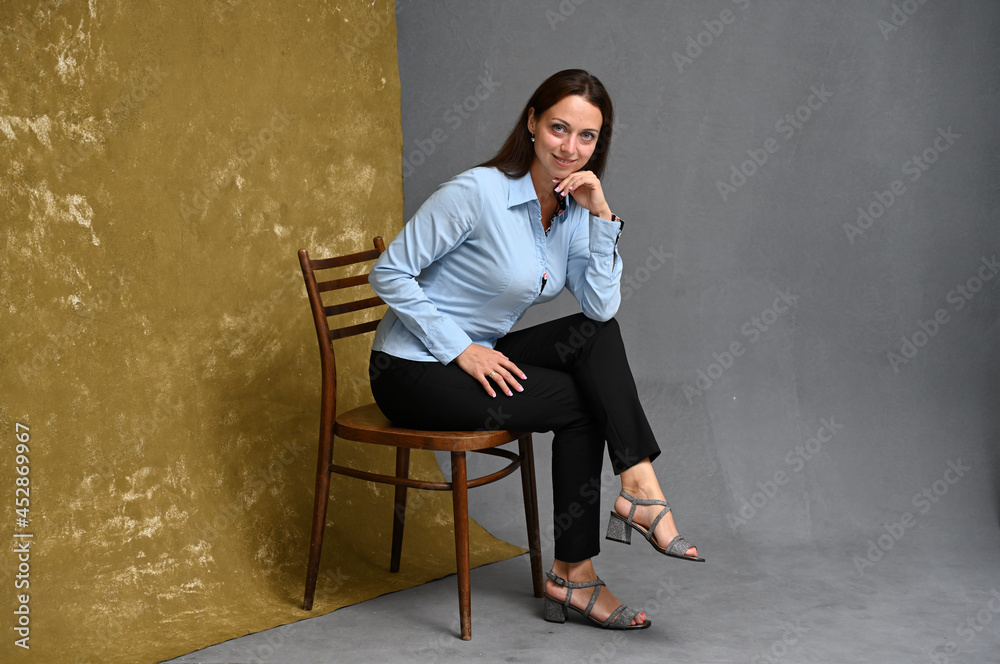 High resolution full length portrait of smiling young woman in blue shirt and trousers posing while sitting on chair against beige and gray background High resolution full length portrait of smiling young woman in blue shirt and trousers posing while sitting on chair against beige and gray background
