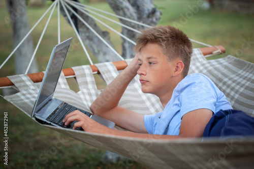 Adorable young teenage man with a laptop lying in hammock in summer garden. Concept distant work, education, freelancer photo