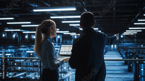 Data Center Male It Specialist and Female e-Business Fintech Startup Manager Talk, Use Laptop. Cloud Computing Server Farm with Two Information Technology Professionals working.