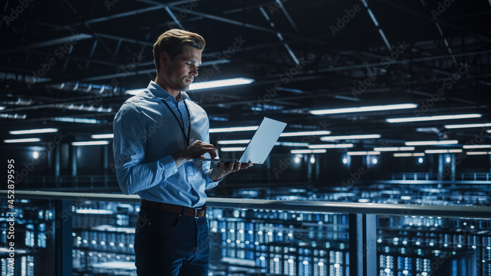 Handsome Smiling IT Specialist Using Laptop Computer in Data Center. Succesful Businessman and e-Business Entrepreneur Overlooking Server Farm Cloud Computing Facility. Handsome Smiling IT Specialist Using Laptop Computer in Data Center. Succesful Businessman and e-Business Entrepreneur Overlooking Server Farm Cloud Computing Facility.
