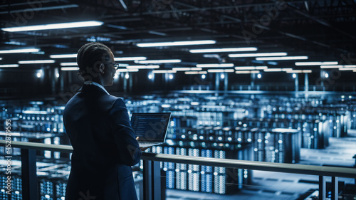 Female IT Specialist Using Laptop Computer in Data Center, Walking on a Bridge Overlooking Big Server Farm Cloud Computing Facility. Businesswoman, e-Business Entrepreneur.