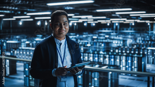 Handsome Smiling IT Specialist Using Tablet Computer in Data Center, Looking at Camera. Succesful Businessman and e-Business Entrepreneur Overlooking Server Farm Cloud Computing Facility.