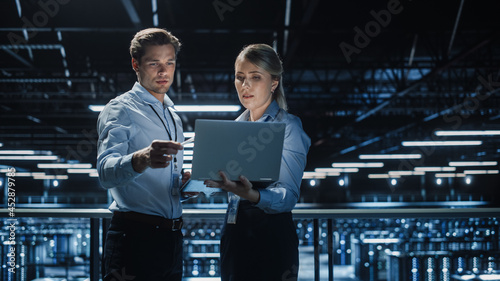 Data Center Female e-Business Enrepreneur and Male IT Specialist talk, Use Laptop. Two Information Technology Professionals on Bridge Overlooking Big Cloud Computing Server Farm. photo