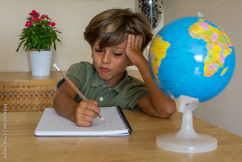 Little Boy In The Living Room Doing Homework. Little Child At Home Drawing With Colored Pencils. He Is A 6 Year Old Caucasian Boy. On The Table He Has A Colored Pencils, A Book And A Globe.Education Little Boy In The Living Room Doing Homework. Little Child At Home Drawing With Colored Pencils. He Is A 6 Year Old Caucasian Boy. On The Table He Has A Colored Pencils, A Book And A Globe.Education