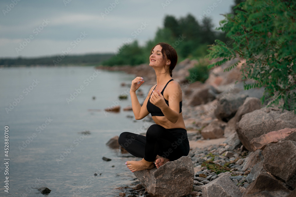 Woman in black sport suit is doing meditation, yoga pose (asana), work out outside near beautiful river and beautiful landscape with wild nature