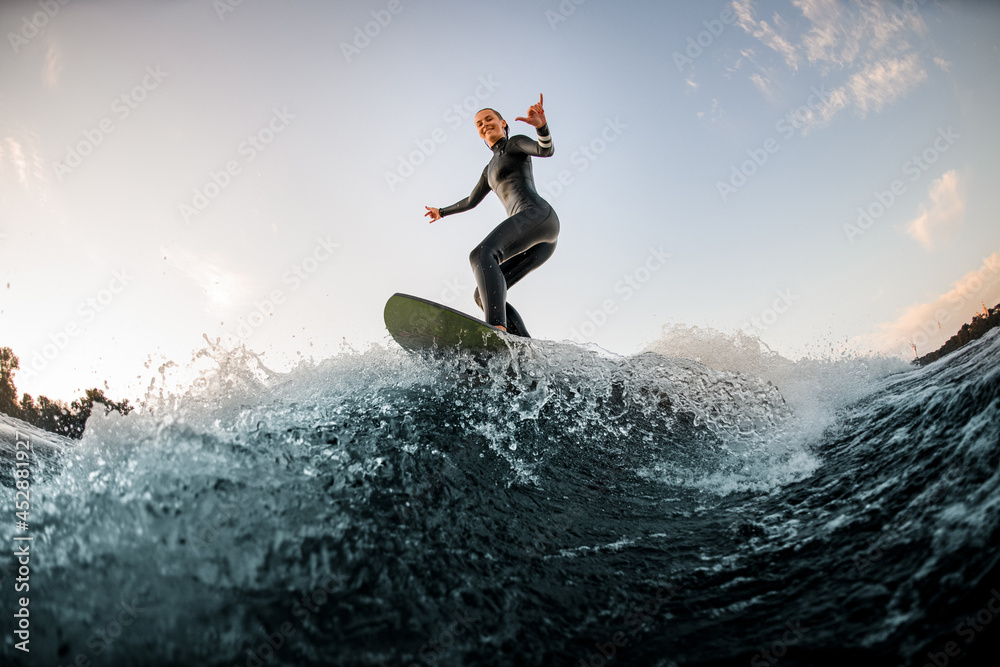 happy woman in wetsuit wakesurfing on the board and riding down the river wave and show hand gesture