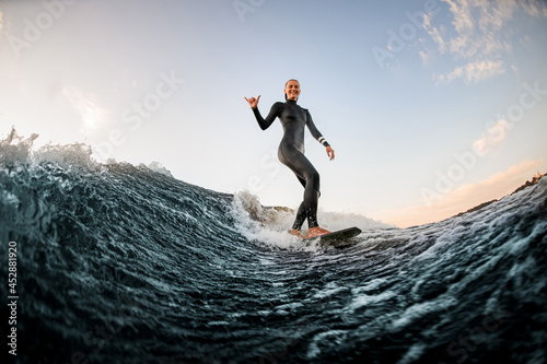 woman in wetsuit wakesurfing on the board and riding down the river wave and show hand gesture