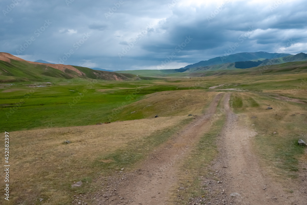 Gravel road on green mountain plateau with mountains on background. Assy plateau, Almaty region, Kazakhstan.Travel, tourism in Kazakhstan concept. Nature landscape.