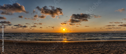 panorama of a colorful beach sunset with the sun dropping into the ocean under an expressive sky