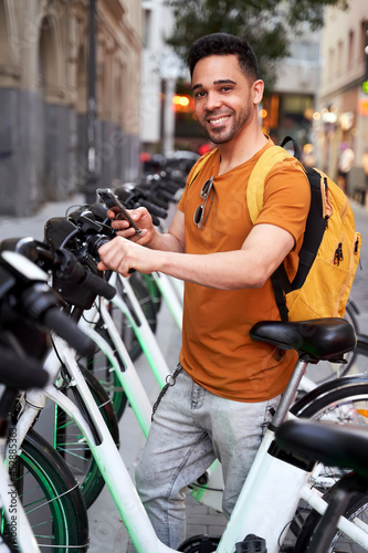 Positive young Latino guy looking at camera stands by the parking lot with a rental bike and uses a smartphone with a smile on his face. Green transportation concept
