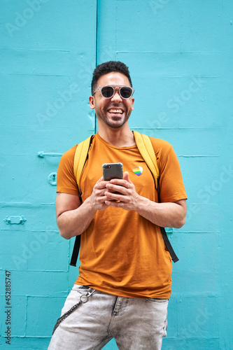 Cheerful young Latino man looking at camera with backpack typing on his cell phone walking down the street, wearing orange t-shirt with lgtbi sticker and sunglasses.