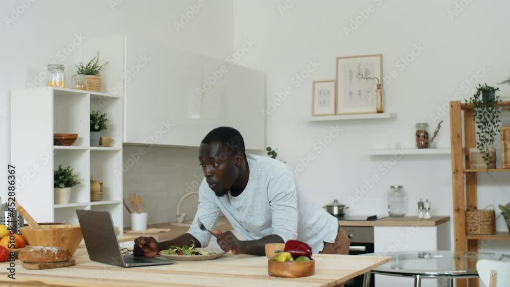 Young Afro-American man leaning on table in kitchen, eating vegetable salad and using laptop Young Afro-American man leaning on table in kitchen, eating vegetable salad and using laptop