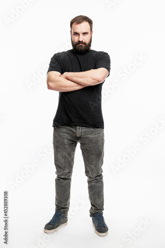 A man with a beard is standing, arms crossed over his chest. Young man in jeans and a black T-shirt. Full height. White background. Vertical.